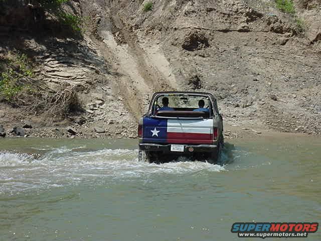 SUV with Texas flag paint driving through shallow water near a dirt embankment