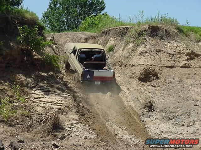 Tan 1978 Ford Bronco driving down a dirt hill, Texas flag on tailgate