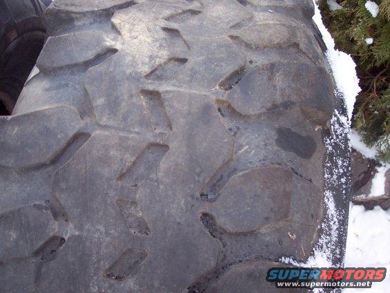 Close-up of a large, aggressive tire tread pattern with snow on the sidewall and a bush in the background. Supermotors.net logo visible