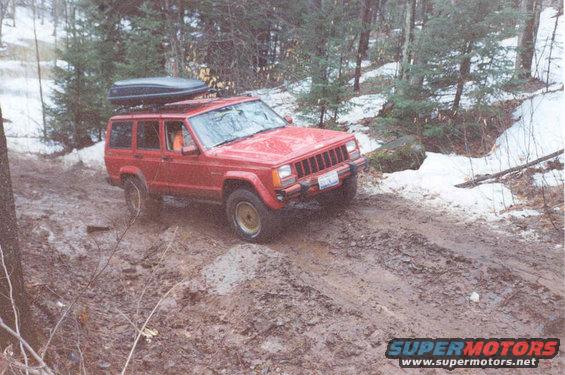 Red Jeep Cherokee XJ with a roof cargo box driving on a muddy, wooded trail with snow patches
