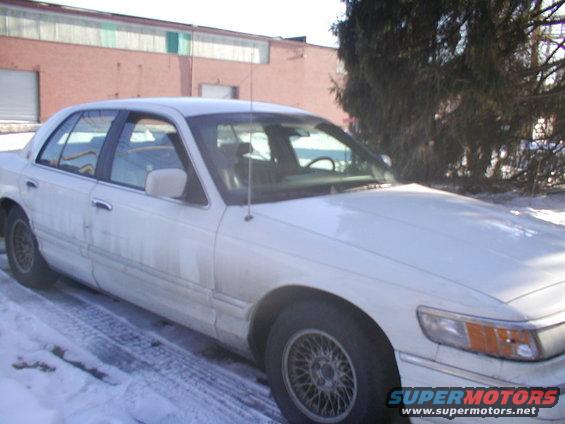 White sedan parked in snow, front quarter view. Building and trees in background