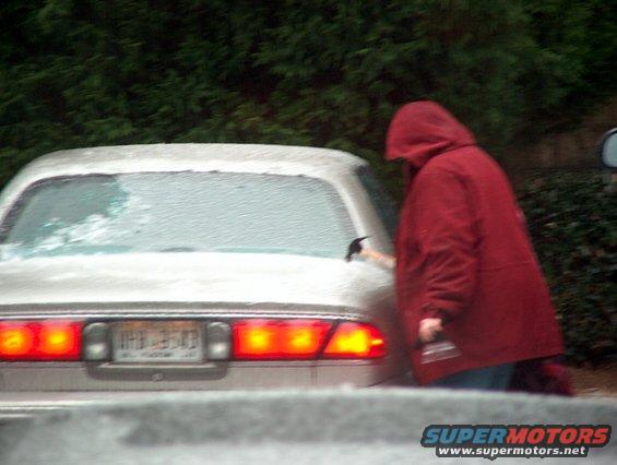1998 Ford Taurus sedan, rear view, light color, with a person in a red hooded jacket scraping ice off the rear window