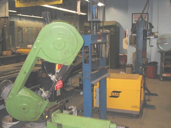 Industrial workshop scene with a green band saw, blue hydraulic press, and yellow ESAB welding machine