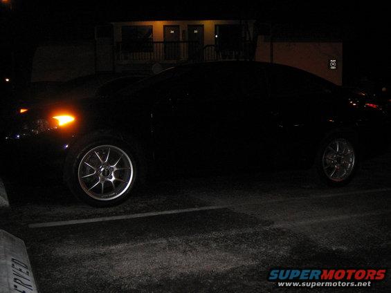 Dark sedan parked at night, showing its side profile and front wheel with amber turn signal illuminated