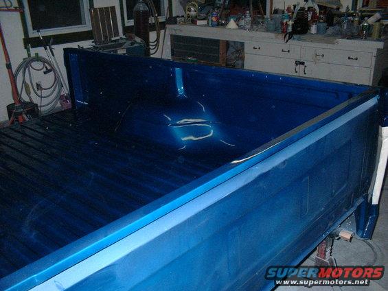 Blue pickup truck bed, showing ribbed floor and side panels. Truck is on jack stands in a garage