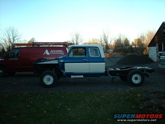 Truck cab and chassis, missing engine and bed. Red van parked nearby
