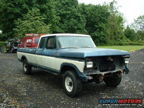 Blue and white Ford pickup truck, missing grille and headlights, parked on gravel