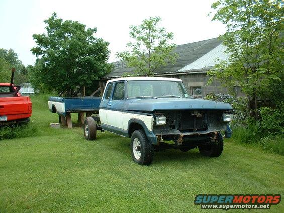 Ford F-Series truck cab and chassis, missing grille and bumper, parked on grass next to a red truck