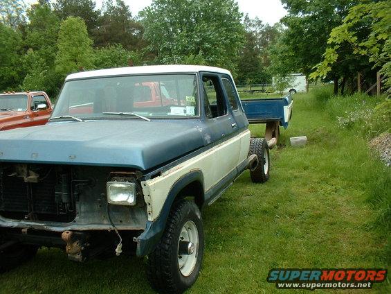 Blue and white Ford F-Series truck, missing grille and headlights, in a grassy yard