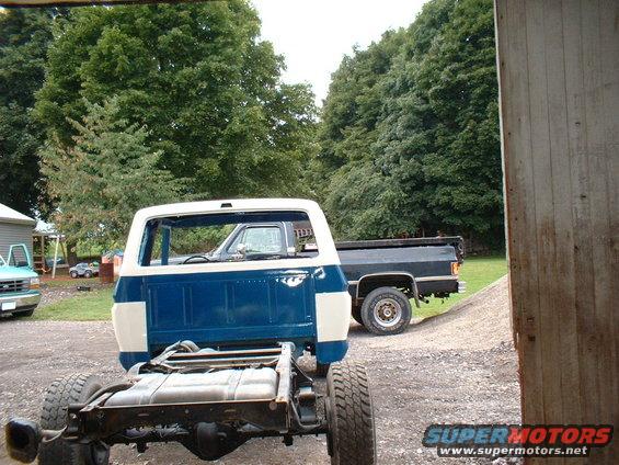 Truck chassis and blue-white cab, rear view. Another dark truck is parked behind it