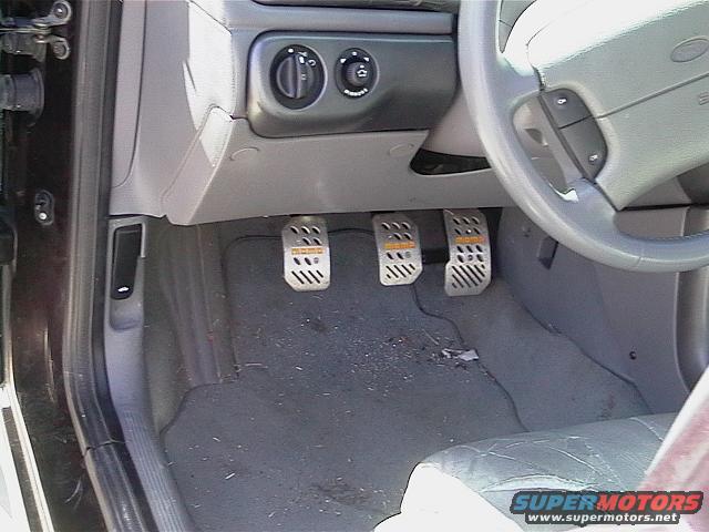 Car interior showing three aftermarket pedals with MOMO branding, steering wheel, and dashboard controls