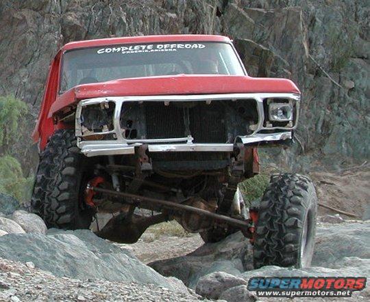 Red Ford truck with large off-road tires navigating rocky terrain. "COMPLETE OFFROAD PHOENIX, ARIZONA" sticker on windshield
