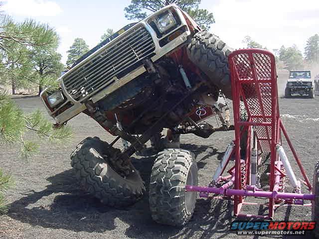Off-road vehicle tilted on its side, front grille and headlights visible, with a red metal structure in the foreground