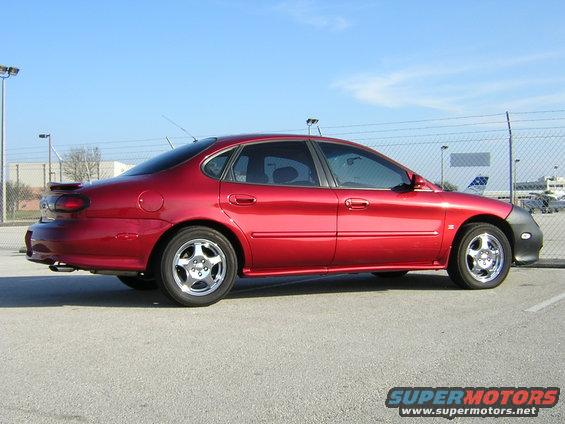 Red sedan parked on asphalt, side view. Has a spoiler and chrome wheels