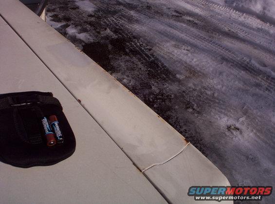 Car hood with a black pouch holding two Duracell batteries, snowy ground with tire tracks