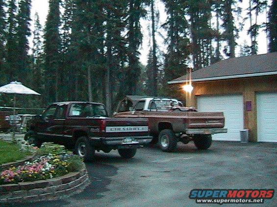 Two Chevrolet pickup trucks parked in front of a garage, one maroon and one brown, with trees in the background