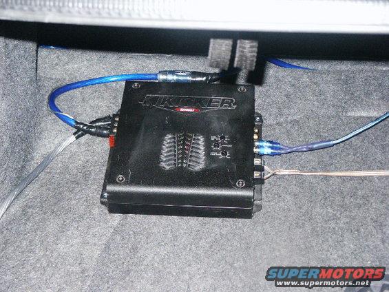 Kicker amplifier with blue power wire and speaker wires connected, mounted in carpeted trunk