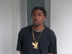 Man with braided hair and a large gold eagle pendant necklace stands in front of a light-colored wall