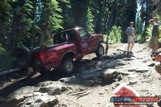 Red 1992 Ford F-150 pickup truck with spare tire on rear, driving on a rocky, wooded trail