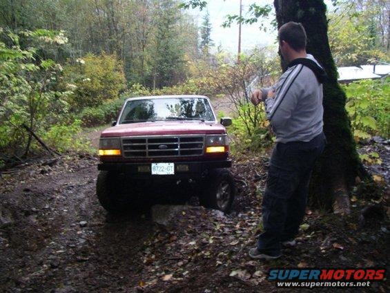 Red 1992 Ford F-150 on a muddy, wooded trail, front view. The truck is angled, with its front left wheel on a rock