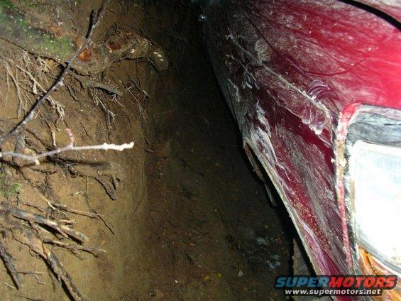 Red vehicle fender and headlight assembly partially buried in dirt with tree roots visible