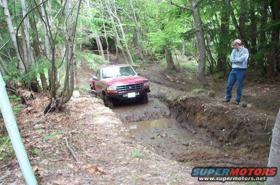 Red pickup truck navigating a muddy ditch on a wooded trail. Guy watches