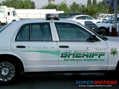 Side view of a white Sheriff's car with green text that reads 'We'll Kick Your Ass' and 'SHERIFF And take your doughnuts too!'