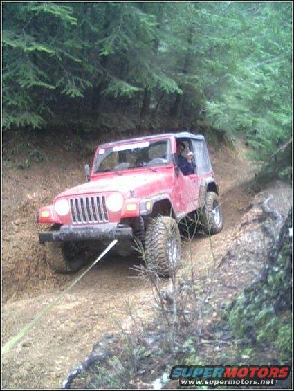 Red 2002 Jeep TJ off-roading on a muddy trail with trees in the background