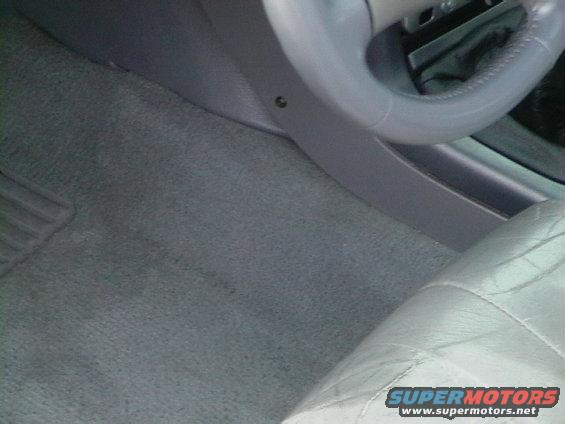 Car interior showing gray carpet, part of a steering wheel, and a quilted seat. Supermotors.net watermark visible