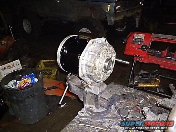 Vehicle transmission part clamped in a vise, with a truck in the background and a bandsaw nearby