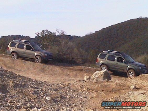 Two green Nissan Xterras driving on a dirt road with hills in the background
