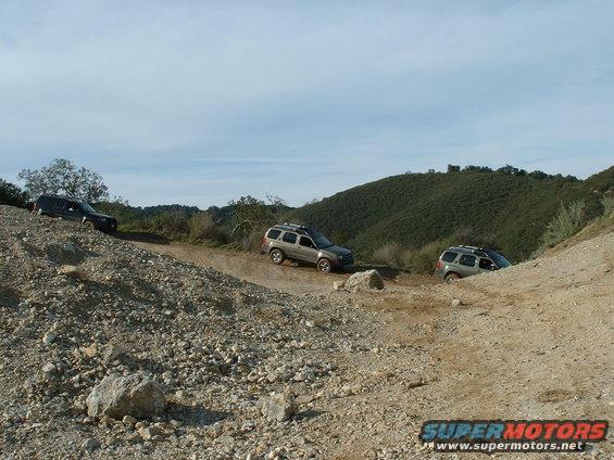 Three SUVs driving on a rocky, off-road trail with green hills in the background