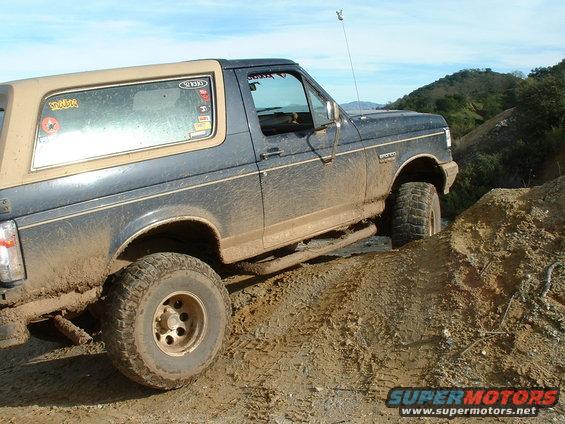 Mud-covered 1991 Ford Bronco driving on a dirt trail with hills in the background