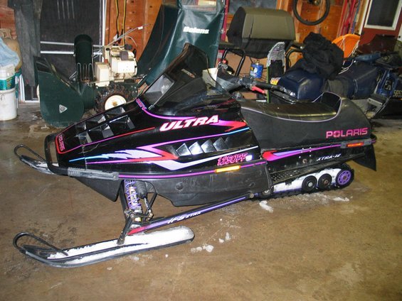 Black Polaris snowmobile with purple and pink graphics, parked indoors next to a snowblower