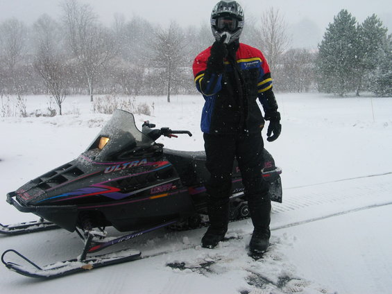 Person in snow gear stands next to a snowmobile in a snowy landscape