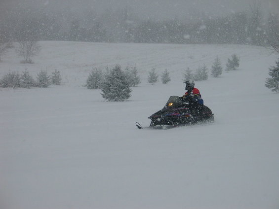 Person riding snowmobile through snowy field with trees in background
