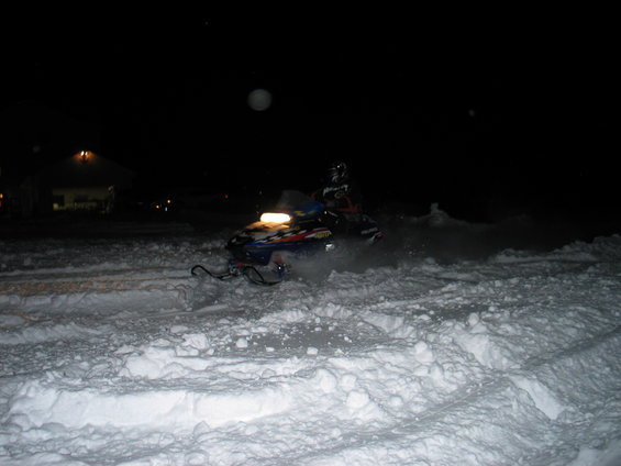 Snowmobile kicking up snow at night, with a building in the background