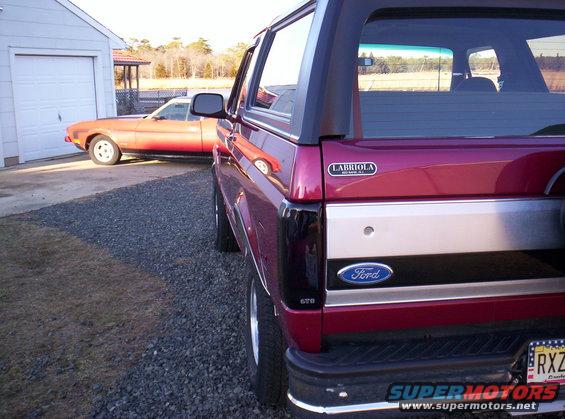 Maroon Ford Bronco rear quarter panel with a silver stripe and Ford logo, parked next to an orange car