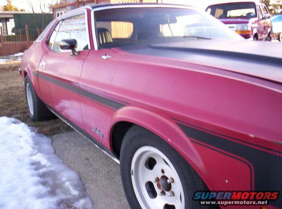 Side view of a red 1971 Ford Mustang Grande with black stripe, parked near snow. Another vehicle is visible in the background