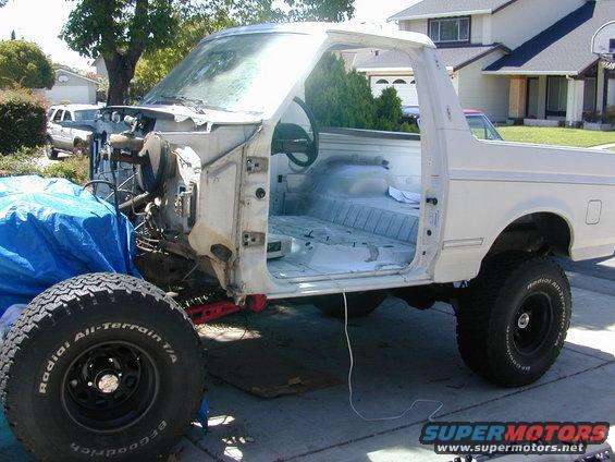 White Ford Bronco body, stripped down, with front end disassembled and interior gutted. A spare tire is visible in the foreground