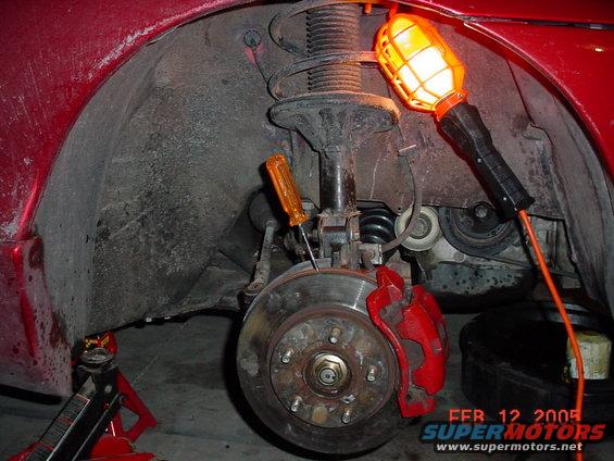 close-up of car's front brake rotor, caliper, and suspension components with a work light and screwdriver