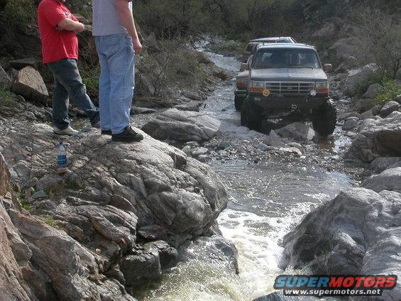 Two people watch as a Ford F-series truck navigates a rocky stream, with another vehicle following behind