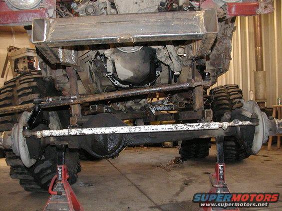 Undercarriage of a heavily modified off-road vehicle on jack stands, showing large tires and suspension components