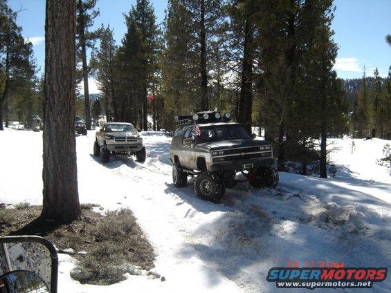 Two lifted trucks being towed in a snowy forest