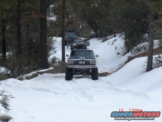 Blue Ford Bronco with big tires driving in snow, other 4x4s behind it