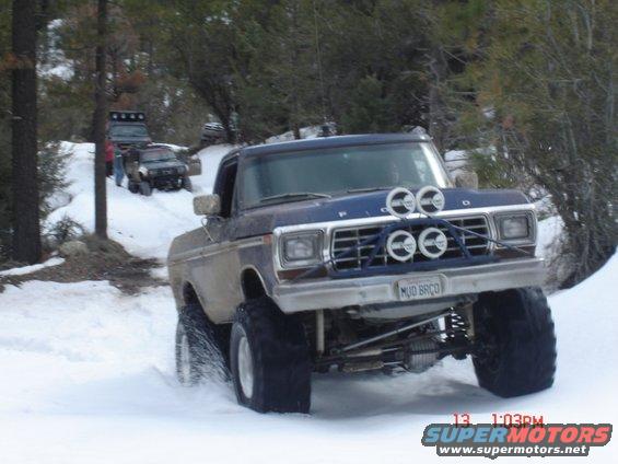 Blue Ford pickup truck with large tires driving in snow, aftermarket lights on grille, license plate reads MUD BRCO