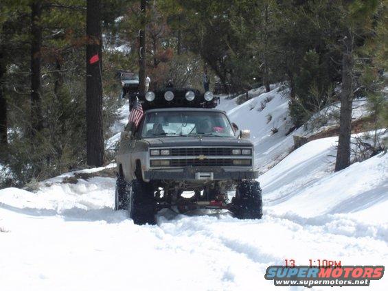 Lifted pickup truck with large tires driving in snow, front view. Modified grille and light bar