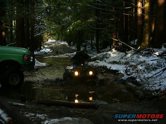 Green truck on left, ATV in muddy forest path with snow