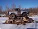 1974 Ford Bronco - A blue 1974 Ford Bronco is stacked on top of a severely damaged gold car in a snowy junkyard setting.