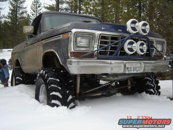 Front view of a lifted blue pickup truck with large tires stuck in snow, custom grille guard and lights